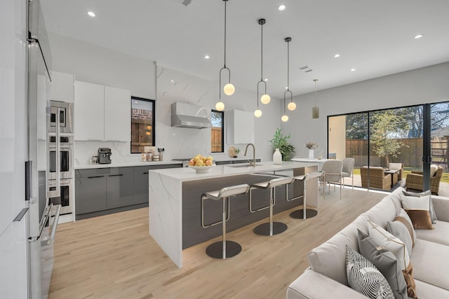 kitchen with decorative light fixtures, a kitchen breakfast bar, gray cabinets, an island with sink, and range hood