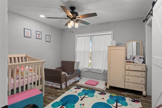 bedroom featuring dark hardwood / wood-style flooring, a barn door, a nursery area, and ceiling fan