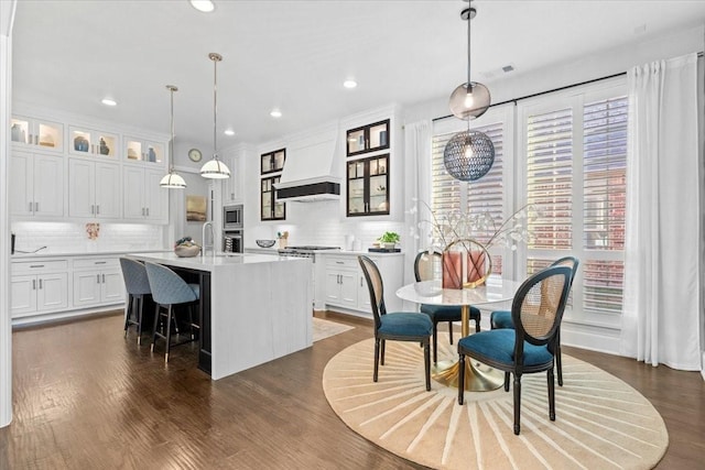 kitchen with pendant lighting, custom exhaust hood, an island with sink, tasteful backsplash, and white cabinetry