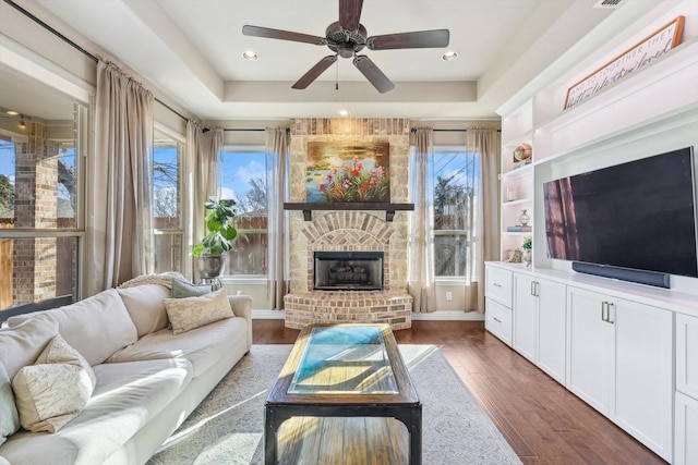 living room with a tray ceiling, ceiling fan, a fireplace, and dark wood-type flooring