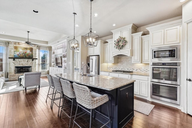 kitchen with stainless steel appliances, a stone fireplace, light stone counters, an island with sink, and a breakfast bar