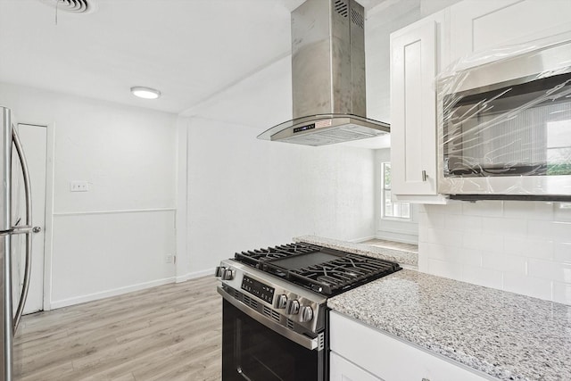 kitchen with wall chimney exhaust hood, light stone counters, appliances with stainless steel finishes, white cabinets, and light wood-type flooring