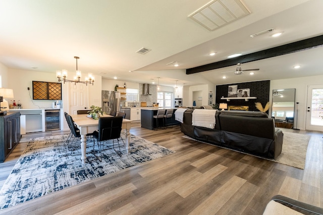 dining area featuring ceiling fan with notable chandelier, lofted ceiling with beams, a brick fireplace, hardwood / wood-style flooring, and beverage cooler