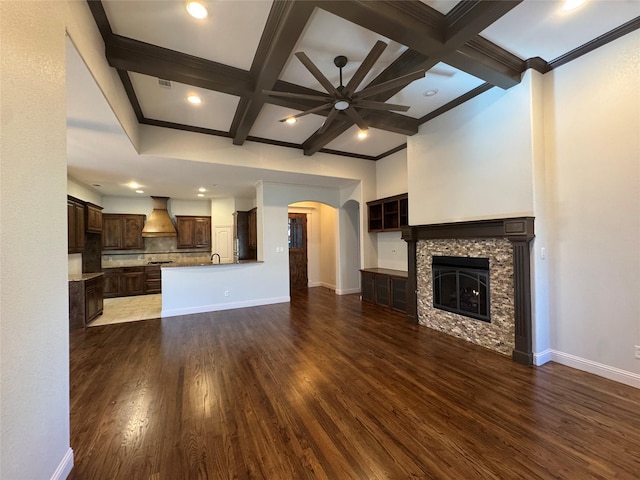 unfurnished living room featuring dark wood-type flooring, coffered ceiling, a stone fireplace, ceiling fan, and beam ceiling