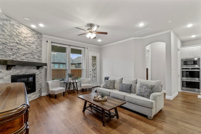 living room featuring ceiling fan, a stone fireplace, wood-type flooring, and ornamental molding