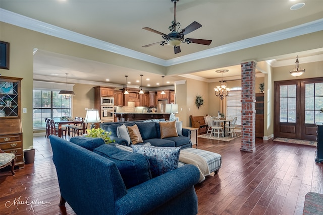 living room with dark wood-type flooring, ceiling fan with notable chandelier, decorative columns, and crown molding