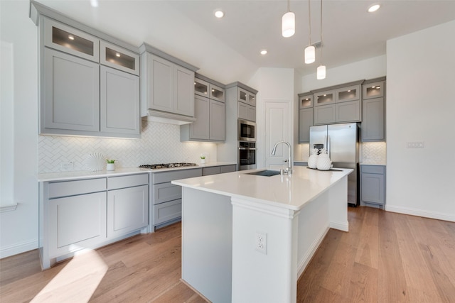 kitchen with light wood-type flooring, stainless steel appliances, sink, hanging light fixtures, and an island with sink