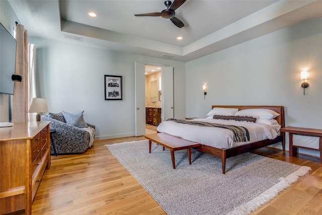 bedroom featuring a raised ceiling, ceiling fan, ensuite bathroom, and light hardwood / wood-style floors