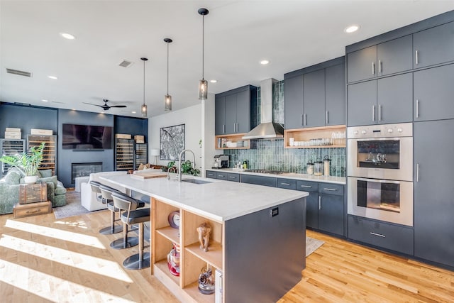 kitchen featuring a kitchen island with sink, wall chimney range hood, hanging light fixtures, decorative backsplash, and stainless steel appliances