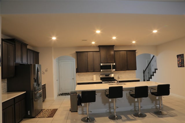 kitchen with dark brown cabinetry, stainless steel appliances, backsplash, a center island with sink, and light tile patterned flooring