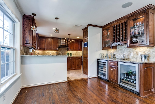 kitchen with light stone countertops, wall chimney range hood, hanging light fixtures, and wine cooler