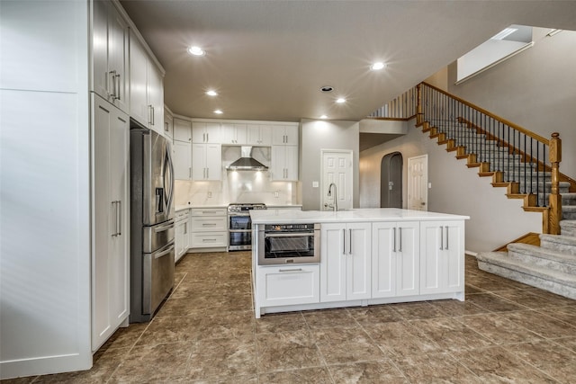 kitchen featuring appliances with stainless steel finishes, white cabinetry, backsplash, wall chimney range hood, and a center island with sink