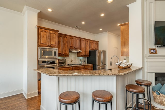 kitchen with light stone counters, under cabinet range hood, a peninsula, appliances with stainless steel finishes, and backsplash