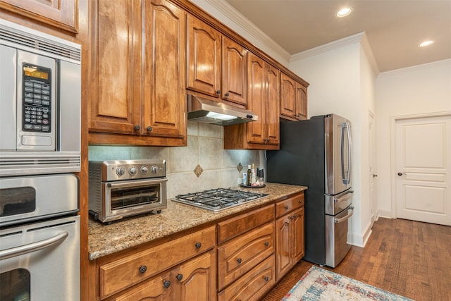 kitchen with light stone counters, appliances with stainless steel finishes, brown cabinets, crown molding, and under cabinet range hood