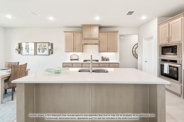 kitchen featuring stainless steel oven, built in microwave, sink, light brown cabinets, and an island with sink