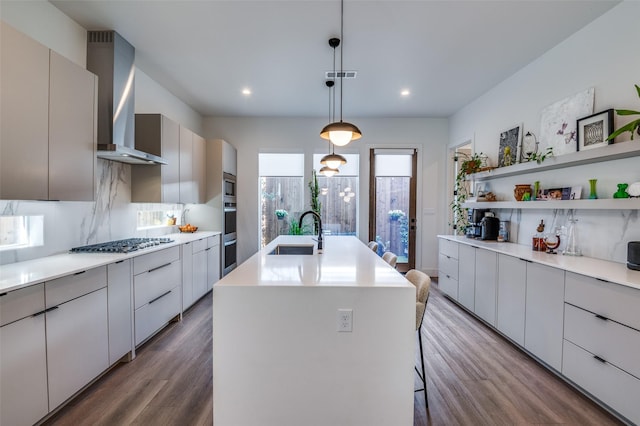 kitchen featuring decorative light fixtures, wall chimney range hood, sink, a kitchen island with sink, and stainless steel appliances