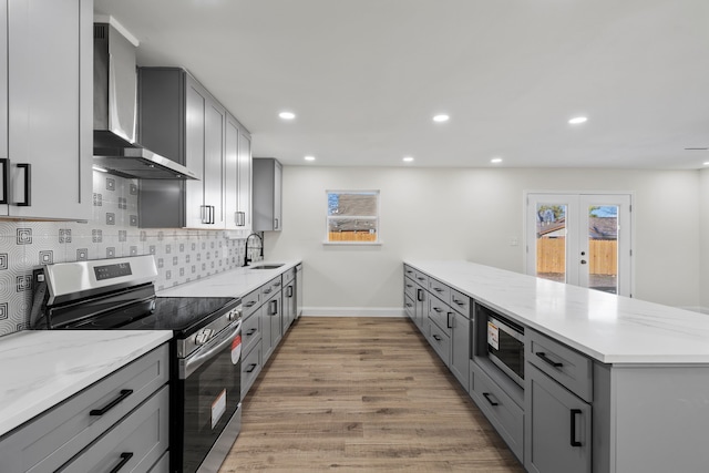 kitchen featuring appliances with stainless steel finishes, gray cabinetry, light wood-type flooring, wall chimney exhaust hood, and light stone counters