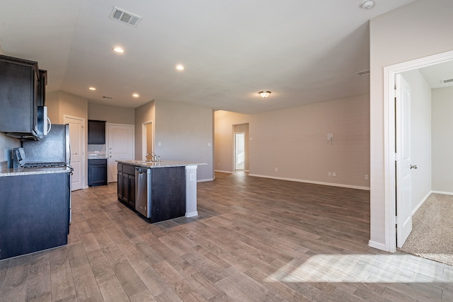 kitchen with light wood-type flooring, light stone counters, stainless steel appliances, sink, and an island with sink