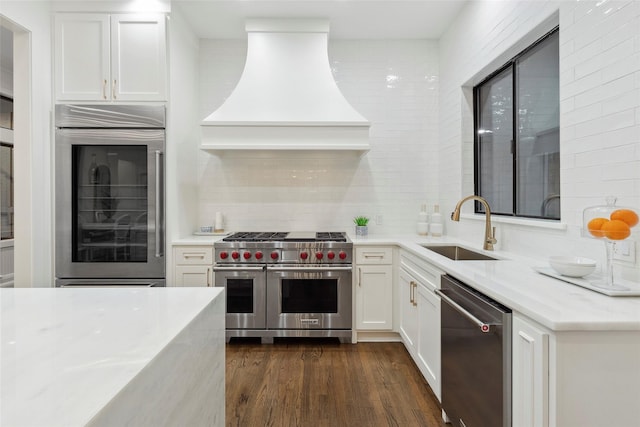 kitchen with light stone counters, custom exhaust hood, high end appliances, sink, and white cabinetry