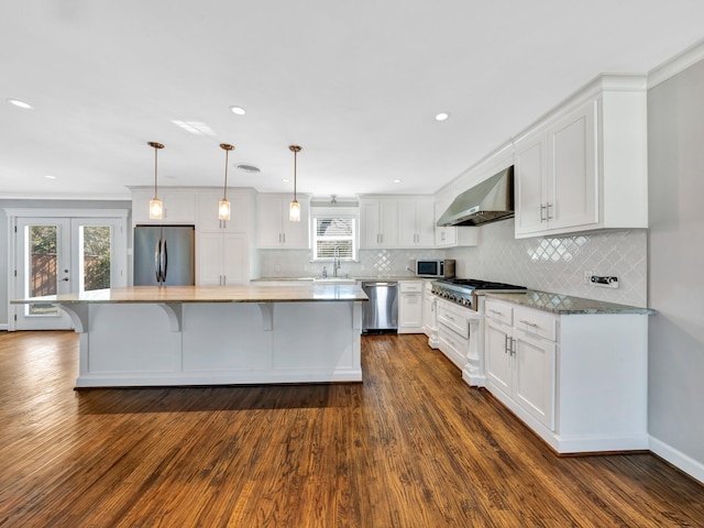 kitchen with wall chimney range hood, decorative light fixtures, a kitchen island, white cabinetry, and stainless steel appliances