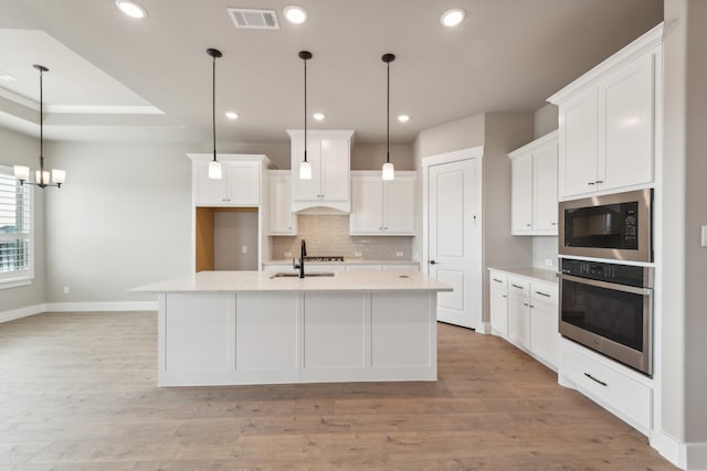 kitchen with stainless steel oven, black microwave, white cabinetry, hanging light fixtures, and an island with sink