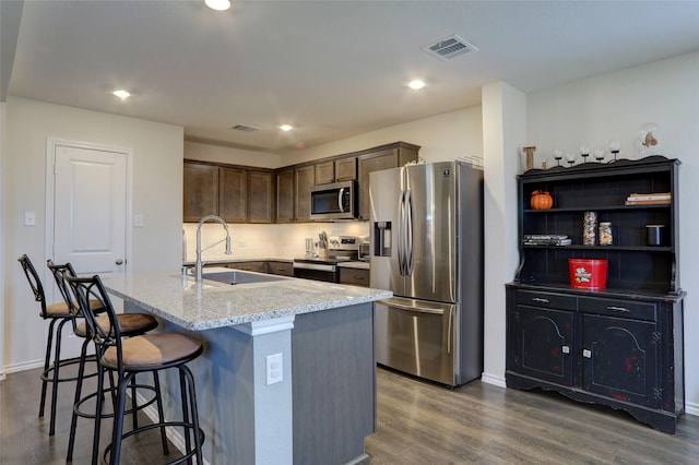 kitchen featuring dark brown cabinetry, sink, light stone counters, stainless steel appliances, and a kitchen island with sink