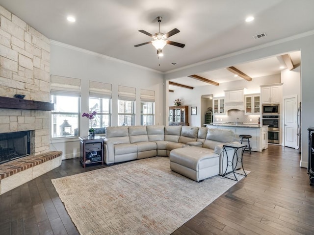 living room with a stone fireplace, ceiling fan, beamed ceiling, and dark wood-type flooring