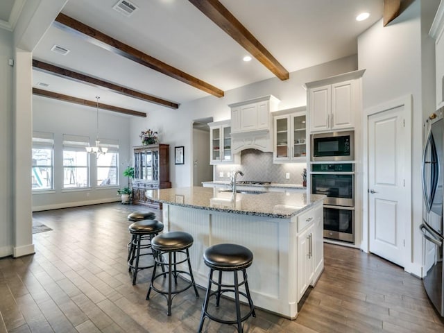 kitchen with appliances with stainless steel finishes, light stone counters, a kitchen island with sink, beamed ceiling, and white cabinets