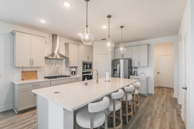 kitchen with wall chimney range hood, stainless steel appliances, hanging light fixtures, and an island with sink