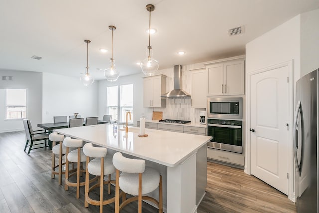 kitchen with wall chimney range hood, hanging light fixtures, an island with sink, tasteful backsplash, and stainless steel appliances