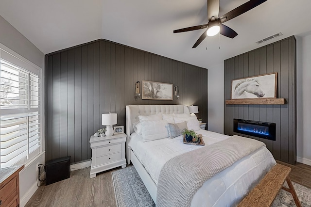 bedroom featuring lofted ceiling, wooden walls, ceiling fan, a large fireplace, and wood-type flooring