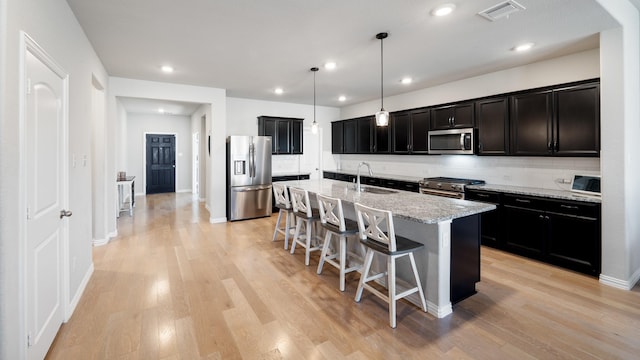 kitchen with light stone countertops, appliances with stainless steel finishes, a kitchen island with sink, sink, and pendant lighting