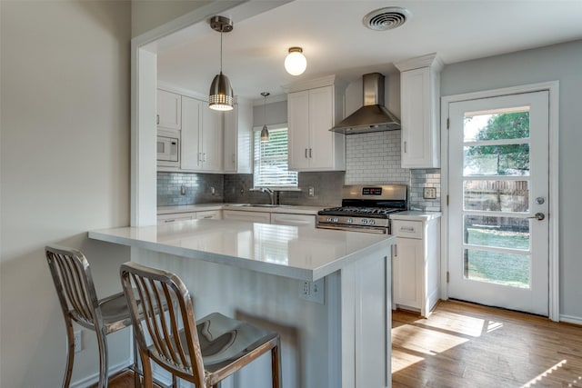 kitchen featuring stainless steel gas range, wall chimney exhaust hood, a kitchen bar, decorative light fixtures, and white cabinetry