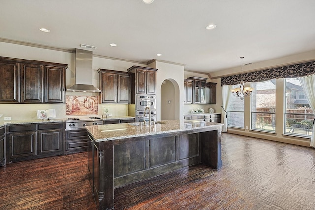 kitchen with wall chimney exhaust hood, an inviting chandelier, a kitchen island with sink, ornamental molding, and dark brown cabinetry