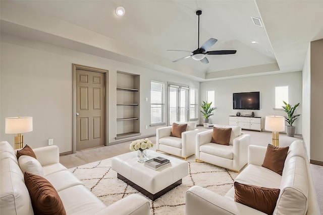 living room featuring ceiling fan, light colored carpet, built in features, and a tray ceiling