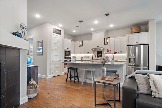 kitchen featuring appliances with stainless steel finishes, an island with sink, pendant lighting, light stone counters, and white cabinetry