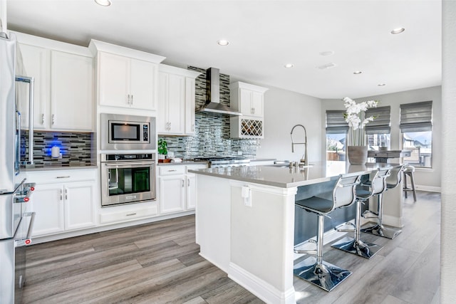 kitchen featuring a center island with sink, a sink, stainless steel appliances, wall chimney range hood, and light wood-type flooring