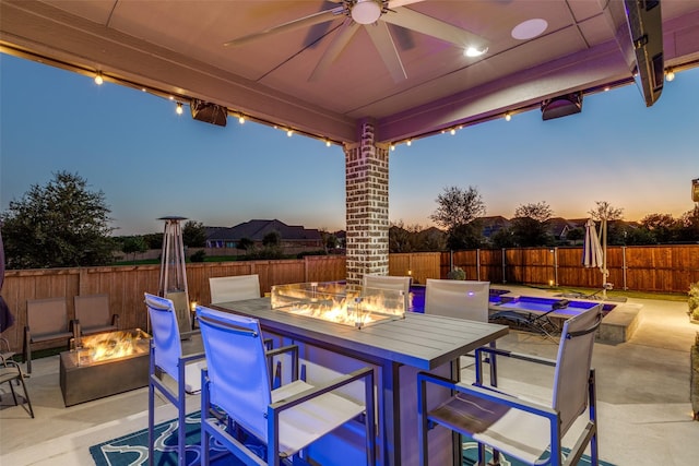 patio terrace at dusk featuring a fenced backyard, a fire pit, outdoor dining space, and a ceiling fan