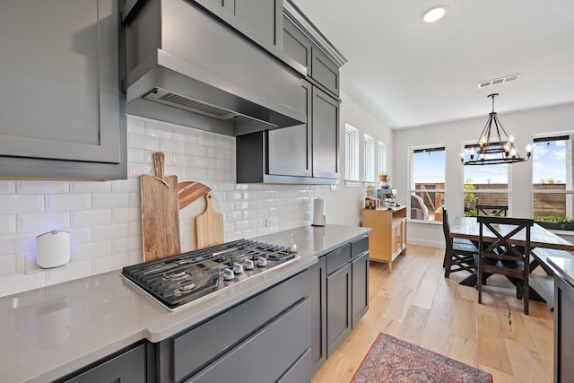 kitchen with a chandelier, gray cabinetry, stainless steel gas stovetop, wall chimney exhaust hood, and tasteful backsplash