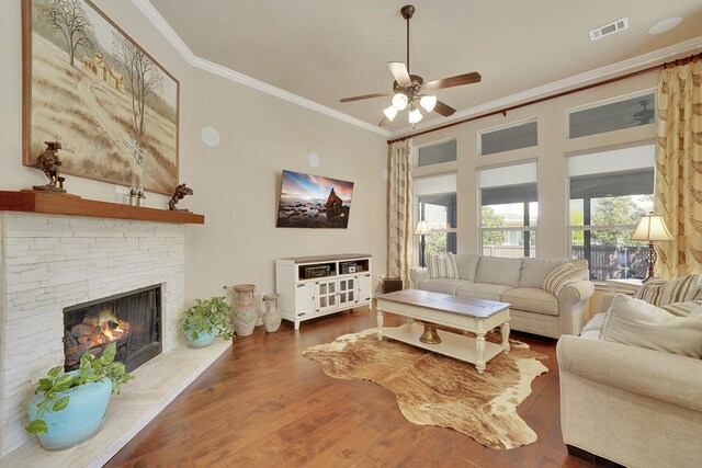 living area featuring a stone fireplace, wood finished floors, a ceiling fan, visible vents, and crown molding