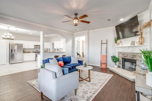 living room featuring ceiling fan with notable chandelier, ornamental molding, light hardwood / wood-style floors, and a stone fireplace