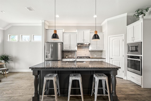 kitchen with decorative backsplash, a kitchen island with sink, white cabinets, appliances with stainless steel finishes, and dark stone countertops