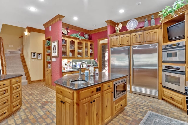 kitchen featuring built in appliances, a kitchen island with sink, crown molding, dark stone countertops, and sink