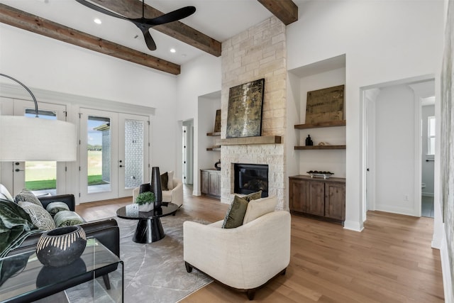 living room featuring beamed ceiling, french doors, ceiling fan, light hardwood / wood-style floors, and a stone fireplace