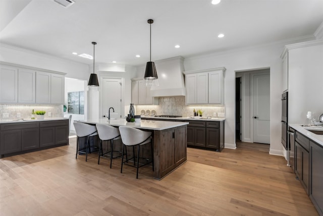 kitchen with hanging light fixtures, an island with sink, custom range hood, stainless steel gas cooktop, and a kitchen breakfast bar