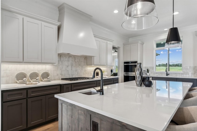 kitchen with hanging light fixtures, a center island with sink, tasteful backsplash, custom range hood, and appliances with stainless steel finishes