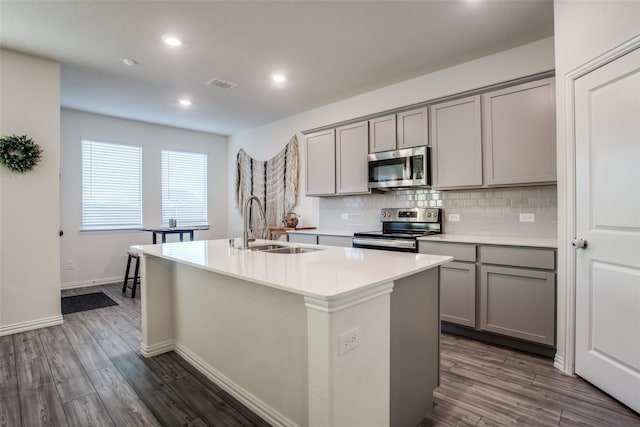 kitchen with dark wood-type flooring, gray cabinets, an island with sink, and appliances with stainless steel finishes