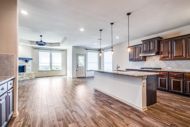 kitchen featuring decorative light fixtures, tasteful backsplash, dark hardwood / wood-style floors, a center island with sink, and a stone fireplace