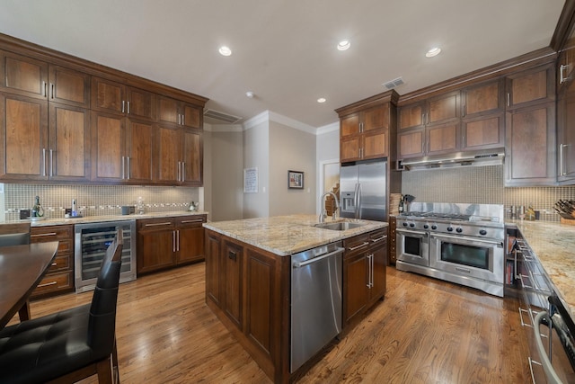 kitchen featuring appliances with stainless steel finishes, backsplash, light stone countertops, a kitchen island with sink, and beverage cooler