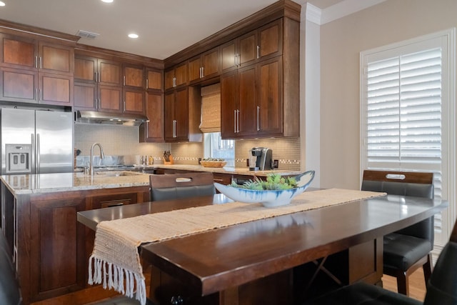 kitchen with tasteful backsplash, sink, light stone counters, a center island with sink, and stainless steel fridge